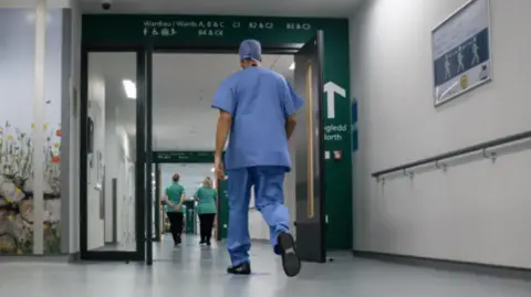 A medical professional wearing blue scrubs walking down a white hospital corridor with a mural of flowers on the left hand side. They are approaching a green open door with ward letters listed above it, with two other medical professionals in front of them. 