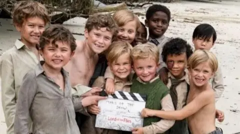 A group of 11 boys with a clipperboard on a beach.