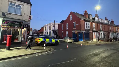 A police vehicle is parked across the end of a terraced street behind a line of police tape.