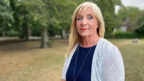A woman with blond hair and a blue cardigan is standing on a grass field in front of trees