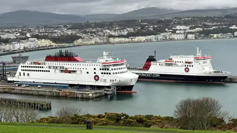 The Manxman and Ben-my-Chree ferries moored in Douglas Harbour. The ferries are painted in the Isle of Man Steam Packet Company colours of white, black and red. buildings on Douglas Promenade can be seen the in background.