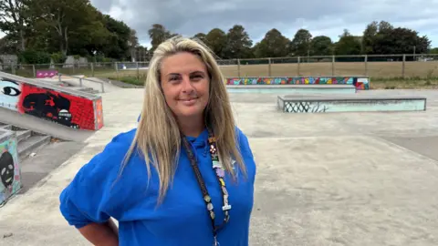 Ally De Cartret - a woman with long blonde hair in a blue hoodie standing in a skate park. 