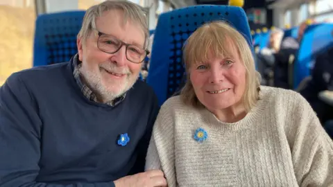 Gordon Weldon sits beside his wife Sue on blue train seats. They are both smiling to the camera and are wearing crocheted blue flowers badges on their jumpers. The badges represent dementia awareness. Gordon has his hand on Sue's arm and is leaning in towards her.  