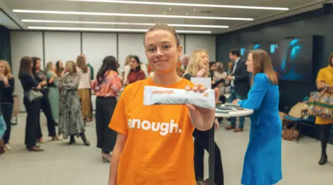 Enough Katie White is standing in the middle of an event room in the foreground with people mingling behind. Katie is smiling and holding an Enough self-swab rape kit towards the camera. She is wearing a bright orange baggy t-shirt that has the word 'enough.' on it in white letters. Her fair hair is tied up tightly.