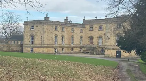 LDRS An exterior shot of Gilling Castle in North Yorkshire, showing the front and a lawn covered in leaves. The grand building has several tall windows and a grand flight of stairs. 