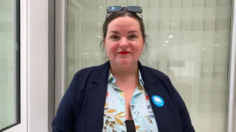 Emma Howgego/BBC Heather Williams standing against a glass window with a door on the right. She has dark brown hair, has her sunglasses perched on the top of her head, is wearing a navy jacket over a floral top and has a blue Conservative badge pinned to her jacket. 