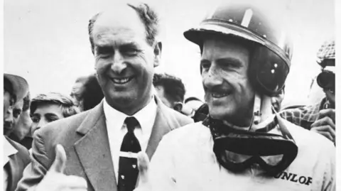 Coleção Hulton-Deutsch/CORBIS/Corbis via Getty Images Uma foto em preto e branco de um homem com cabelo curto nas laterais, vestindo terno e gravata e com os polegares para cima. Ao lado dele está um homem mais jovem, usando capacete e óculos de proteção no pescoço, olhando para o lado e sorrindo. Uma multidão pode ser vista atrás deles.