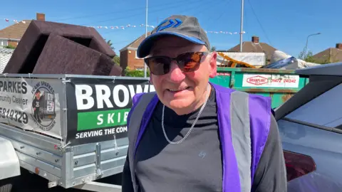 An elderly man wearing a baseball cap, tinted sunglasses and a hi-vis vest over his T-shirt smiles for a photograph.