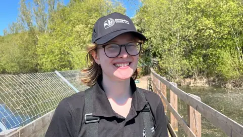 Ellen Knight/BBC Clement pictured looking into the camera and smiling. Clement is wearing a black polo shirt, a black baseball cap, and glasses. She has red shoulder-length hair. They are stood on the board walk in Shrewsbury's Old River Bed, a long wooden bridge. 