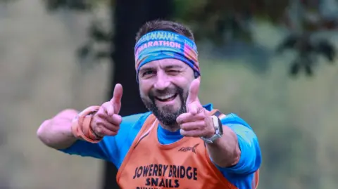 A man with a head band in bright colours wearing a blue T shirt with an orange vest over the top. He is smiling and pointing his fingers at the camera