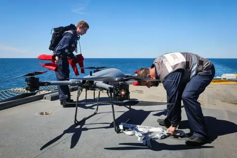 LPhot Bill Spurr Two men wearing Royal Navy boiler suits, attach elements to a Malloy drone on a ship with the sea in the background 