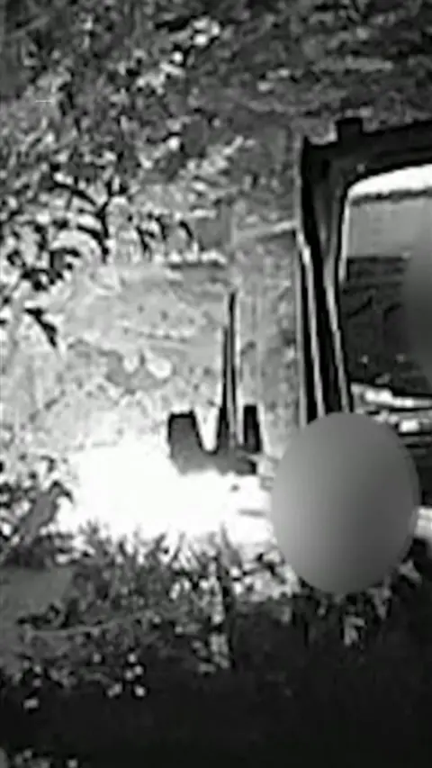 A black and white cctv image. There is a blue over a persons face. To the right of the person is a truck. There are trees with leaves around.