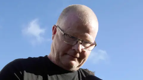 PA Media Richard Satchwell is wearing a black t-shirt and he has a bald head with square glasses on. He is looking down. The sky behind him is bright blue.