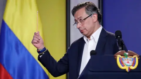 EPA A middle-aged man in a blue jacket and white shirt waves a pencil while standing next to a Colombian flag.