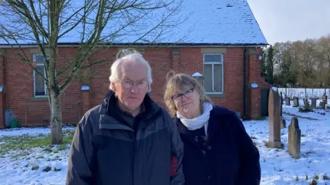 BBC Couple standing in front of a disused chapel in a snowy graveyard, surrounded by headstones.
