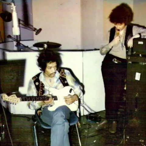 A. N. Other Jim Hendrix is seated and playing a white electric guitar in a recording studio. Another person is standing nearby, leaning on a piece of audio equipment and holding a microphone.