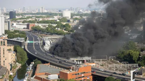 Submitted Thick black smoke billows from beside the elevated A40 Westway flyover at Westbourne Park, with the Grand Union Canal, railway lines and the central London skyline visible behind.