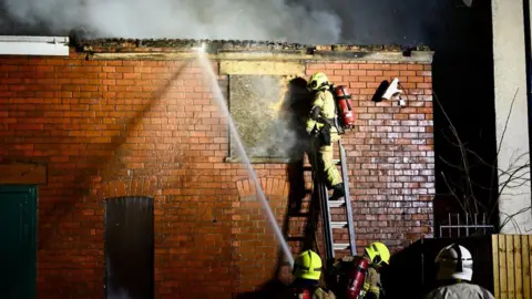 Shot of a firefighter on a ladder tackling the blaze with colleagues below with hoses. There is smoke coming from the red-bricked building and a security camera in view.