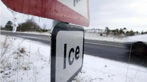 A road sign saying: 'Ice' has frozen raindrops hanging from it. Snow can be seen on the sides of road behind it. 