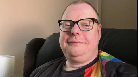SUPPLIED Man with glasses in black t shirt with rainbow shoulder pattern sit on dark sofa against cream wall