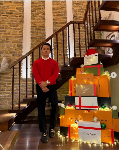 Instagram/sgtthb Saugat Thapa, con un suéter rojo y pantalones negros, se encuentra junto a un árbol de Navidad hecho con cajas de marcas de lujo.