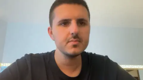 A man with short brown hair sitting in a hotel room looks into a camera