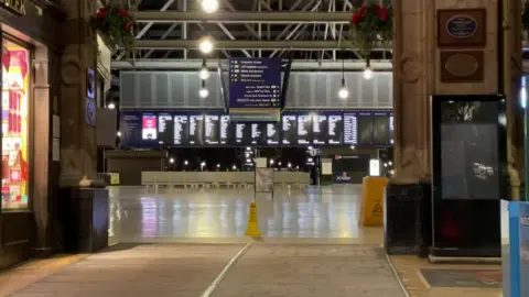 The concourse at Glasgow Central Station. It is empty. A yellow wet floor sign is in the middle. There is a shiny white floor. In the background are screens displaying travel information.