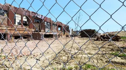 A derelict building behind a wire fence Beside dry soil with rubble and grass in places. Something is stacked in the background, possibly fencing or planks of wood. The day is sunny and the sky is blue. 