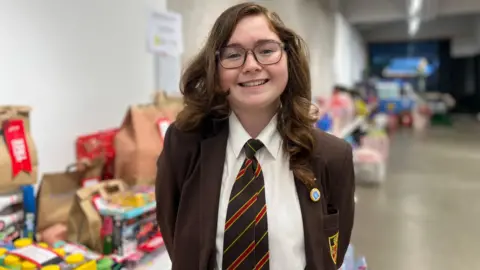Allarna stands in front of a table full of toys and games. She has long, dark wavy hair. She wears a brown school blazer and a brown and orange striped tie