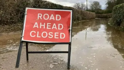 BBC A red sign reads road ahead closed on a country, flooded road