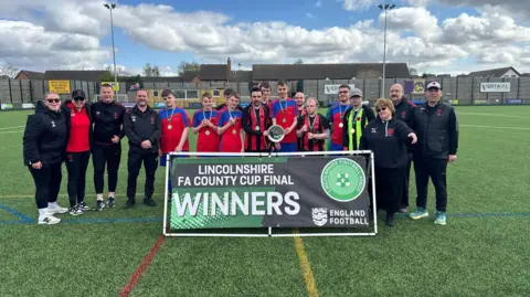Barton Inclusive Football Club A line-up of players and coaching staff standing behind a placard which reads "Lincolnshire FA County Cup Final Winners".