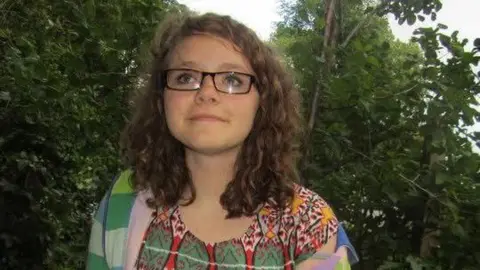 Tessa Marshall Tessa, who has rectangle framed glasses and dark curly hair. She is wearing a colourful top with green, red, and yellow patterns on it, and a green and blue striped cardigan. Trees can be seen behind her. 