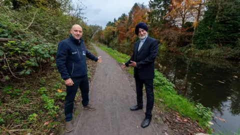 City of Wolverhampton Two men stand on a newly-resurfaced path by the side of a canal. One man is wearing a suit and a turban, the other is wearing a navy blue jacket and navy blue trousers and brown shoes.