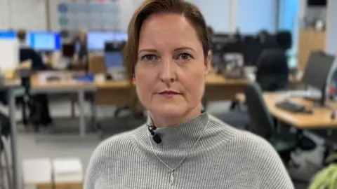 Alison Hernandez has brown hair pulled back from her face. She is wearing a grey jumper and standing near a sign which reads PCC Office of the Police and Crime Commissioner for Devon and Cornwall.