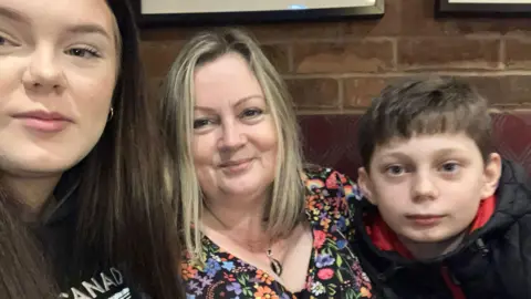 A young girl with dark hair on the left, a woman with blond hair wearing a flowery top in the middle and a young boy on the right wearing a dark coat. All are sat down with a brick wall backdrop behind them