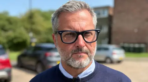Craig is wearing a blue jumper and shirt and glasses. He has grey hair and a beard. He is stood outside near some cars.