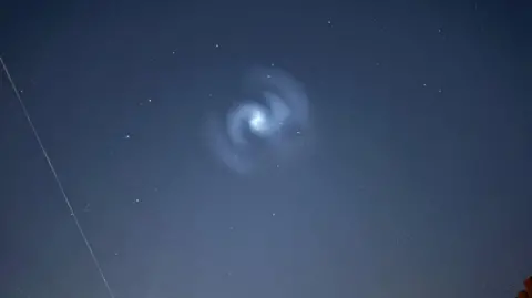 A dark night sky with a spiral light in the middle. There are several stars surrounding the light.