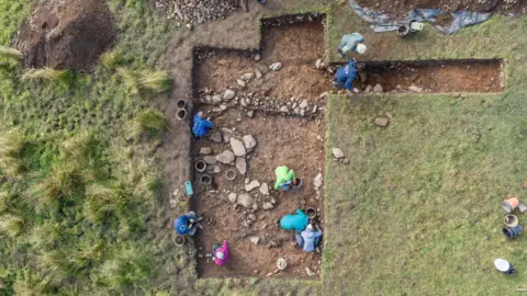 An aerial image of the excavation site people kneeling down with buckets and spades inside two rectangular shallow pits with exposed soil and stones