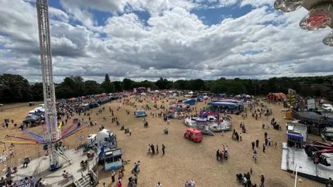 A view looking down at a festival in a park, showing crowds of people, fairground rides, and various stalls