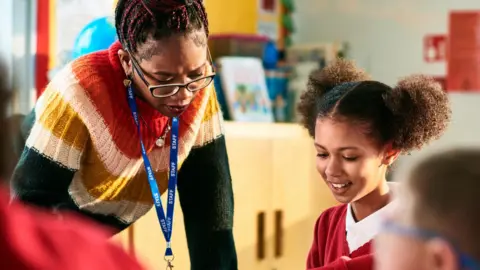 Getty Images Cheerful school girl showing her book to her teacher who is leaning over the desk and concentrating
