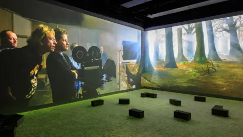 Justin Sutcliffe A projection screen shows four men standing behind a studio camera looking at a forest. In front of the screen is a seating area. 