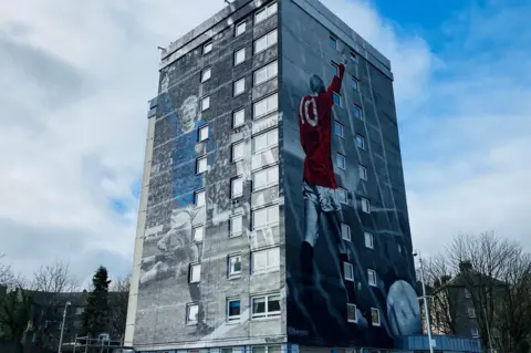 BBC High-rise building with murals of late footballer Denis Law with his arm aloft, one in a red shirt, one in a blue Scotland shirt.