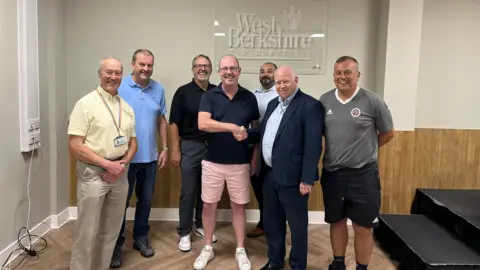 West Berkshire Council Representatives of West Berkshire Council and NCFG meeting in front of a West Berkshire Council sign. They are Cllr Nigel Foot from West Berkshire Council - yellow shirt with stone-coloured chinos; John Stewart from NCFG - blue T-shirt and blue trousers with trainers; Cllr Vaughan Miller from Newbury Town Council - wearing a black T-shirt, grey trousers and white trainers; Lee McDougall from NCFG, wearing pink shorts, white trainers and a black T-shirt; Nick Robbins from NCFG - wearing a blue open-necked shirt, who has a beard and short dark hair; Jeff Brooks, the council leader, wearing a dark suit and a white shirt, and Jason Braidwood - who is wearing a grey Adidas T-shirt and dark Adidas shorts