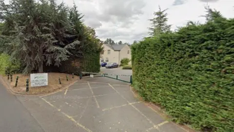 Google A section of a cream coloured building with a grey roof can be seen in the gap between two connifer bushes. There is a concrete driveway leading up to the house with yellow hatching in front of a small metal gate. 