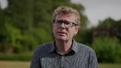 A man in a grey floral shirt with glasses and curly brown hair
