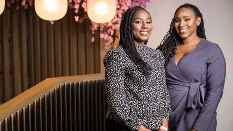 Two black women with long hair stand next to each other and are smiling at the camera while looking at it. One is wearing a dotty top and the other is wearing a purple dress. To the left of them is a bannister, lights hanging from the ceiling and pink flowers. 