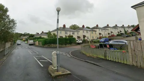 St Thomas' Crescent at the junction with Sycamore Avenue. A traffic island stands in the middle of the road and speed bumps can be seen further down the road. A road of semi-detached white houses line Sycamore Avenue to the right.
