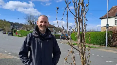 Darren Parkinson A man in a waterproof jacket with his hands in his pockets stands next to a small tree on a quiet residential street. He is smiling at the camera. It is a sunny day.