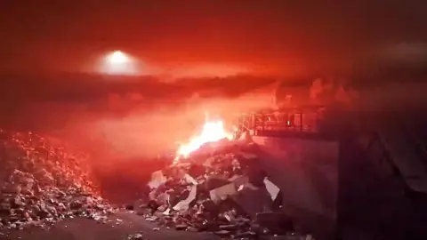 FCC ENVIRONMENT The image appears to be from a CCTV camera and shows piles of rubbish in the foreground of the picture with flames on top of one of the piles. Much of the picture is covered in a red haze with some smoke visible. What appears to be a light can be seen to the top left of the picture through the haze.