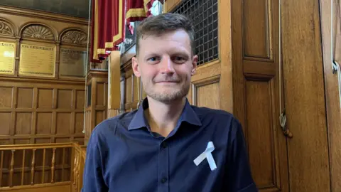 A man with short brown hair and stubble is wearing a navy short sleeved shirt and a white ribbon pinned to his shoulder. He is standing inside a town hall chamber